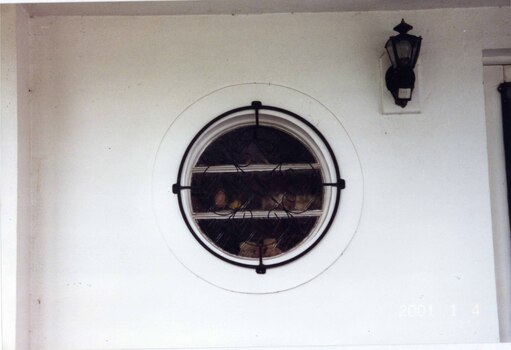 White wall containing a round window circled by a black metal ring and lacing over the window.  Shelves visible behind the glass with 3 rows of objects.  Small black metal and glass lamp above and to right of the window.