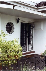 Entrance porch with round window circled by black metal and lace-like grating, which matches the decoration of the black double outer doors (in front of double glass doors) to the right of the window and the black metal decorated fence along the edge of the porch landing.  Black metal and glass light to the left of the doors and a white box with small round handle to the right of the doors.  Bushy pot plants on the left of the porch with some garden plants in front of the porch.