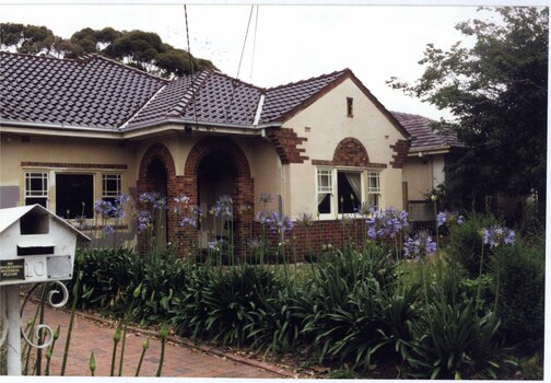 Partial front view of a cream house with brown brick features around the double-arched porch entrance, windows, eaves and bottom of the walls.  White decorative metal mailbox with the number 10 on it in front of a mixed brown paved drive lined with tall plants with purple flowers.  Lawn is behind that in front of the house.  Next door house just visible to the right side and tall trees behind and right front.