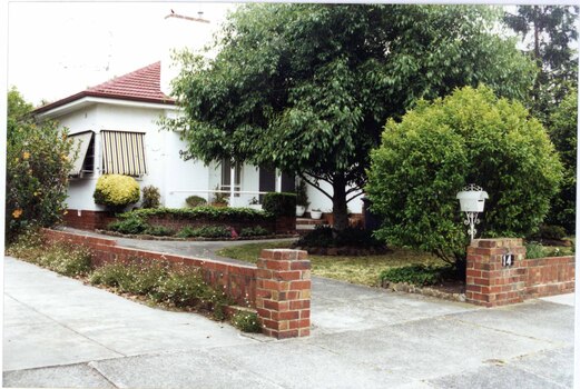 Partial side and front view of white house with brown brick features, double glass front doors and striped awnings.  Low brown brick side and front fences with white letterbox.  Large tree in a round garden bed in the lawn with other garden beds plus large and small plants in the garden with drive on the left.