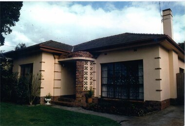 Front view of very light brown house with mixed browns brick features and a porch with a decorated curved side.  Garden beds in front of house include larger plants on left side and smaller side on right side.