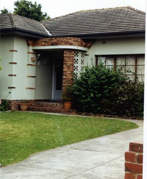 Front view of light green house with mixed browns brick features and a porch with a decorated curved side. Garden bed in front right of house has well-established plants.
