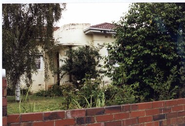 Partial front view of cream house with a porch with curved wall and columns in a garden of well-established trees and shrubs with a lawn.  Brown brick front fence with part of its pillar showing to the left.