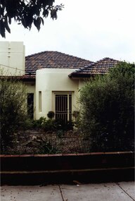 Partial front view of white house with a porch having a curved wall, long narrow window and full height metal gate entry.  Broad white chimney on left of roof.  Behind a brown brick fence is a garden of tanbark and well-established shrubs.