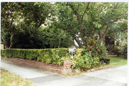 Obscured front view of white house with brown brick edging around the porch door entrance.  Brown brick fence with trimmed hedge above it, with a well established garden of trees, shrubs and lawn all to the left of the drive.