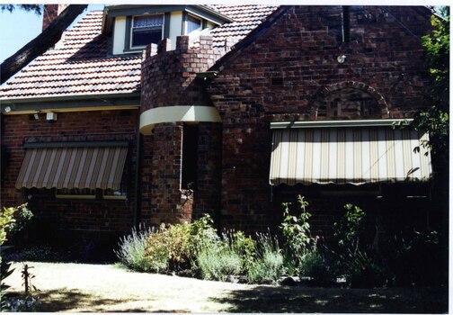 Partial front view of variegated brown brick house with attic window jutting out of its steep tiled roof.  Round tower-like porch entry with long narrow open window.  Striped awnings on the windows above the garden beds of small plants with a path to the front porch.