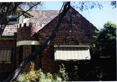 Front view of the right of this variegated brown brick house including cream features with a tower-like porch, attic window and striped awnings over the windows. Garden bed of small plants with medium sized tree to the right.  A large tree branch is in front of the house.  