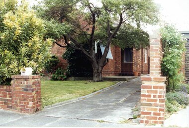 Partial front and side view of a variegated brown brick house with an open doorway square porch.  Striped awning on one of the windows.  Well established tree and garden to the left of the concrete drive.  Low matching brown brick front wall with white letterbox on the fence pillar and high brown brick side fence.