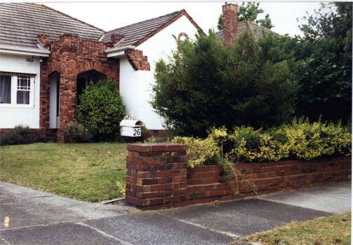 Partial view of white house with variegated brown brick features including the square double arched porch. Matching brown brick low fence with drive to the left and the central lawn has smaller plants around the house plus larger ones on the fence line