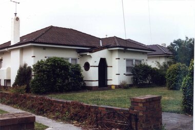 Side and front view of a white house with brown brick features and matching brick low side fence along the drives in the foreground.  Arched entrance to the porch which has a round window on the side.  Chimney has a circle decoration facing the front.  Well-established bushes lining the house and front with an open lawn area. 