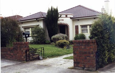 Front view of white house with brown brick features, especially decorative around the wide arch of the porch with a long brick panel to the right of it.  It has 2 tall chimneys, striped awnings, established shrubs around its lawn, a matching brown brick low fence with pillars each side of the drive and a black metal letterbox on a pole over the left pillar, plus a black metal gate on one side.