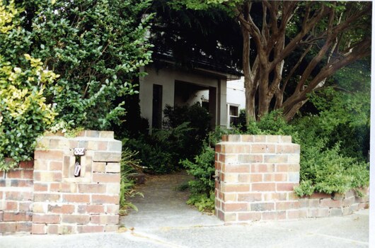 Beige house with a large porch of square arches on 2 sides.  It is mostly hidden behind the tall shrubs and trees with a concrete path leading from the low brick fence to the entrance.