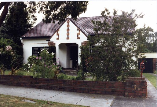 White house with brown brick features alongside matching low brick fence with brown metal letterbox and rear garage with matching brick feature wall to right.  Wide arched porch with black framing the windows on either side of it plus black framed entrance to garage, down the right-side  drive.