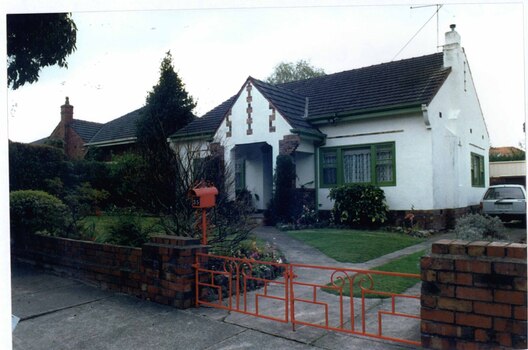 Front and right side view of white house with brown brick features alongside matching low brick fence with brown metal letterbox and matching metal gates.  Also a rear garage with matching brick feature wall to right. Wide arched porch with green framing the windows on either side of it plus the gutters and drain pipes.  Well-established garden and left neighbour's house partially visible.