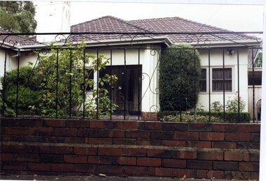 Partial front view through black metal lace fence of a white house with a large porch with matching black metal lace door with a column to the right.  Large chimney and a well-established garden.