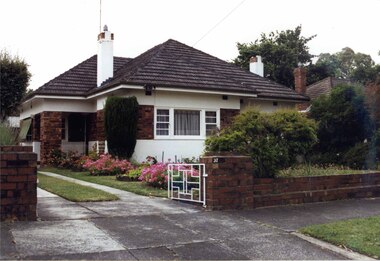 Partial side and front view of rendered house with large band of brown bricks wrapping around the house.  Large open porch with brown brick pillar, two chimneys with matching brick tops and matching brick low fence with pillars on either side of the drive and a white metal fence.  Garden has 4 circular beds along the drive on the side of the lawn.  Roof and chimney of right neighbour is just visible.