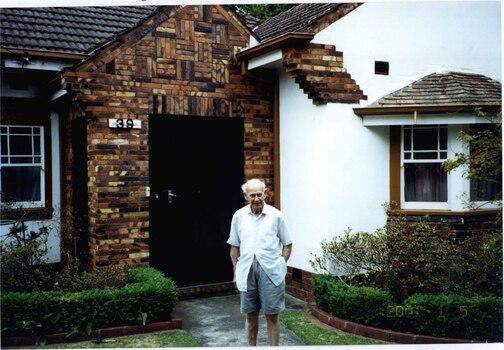 Partial front view of variegated brown brick large porch with black metal door for white house.  Well-established garden and elderly man standing on concrete path in front of the house.