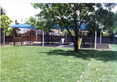 Several items of playground equipment under a large blue roof.  Grass and a tree in front of the playground and a fence and houses behind it.