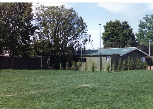 Large grassed area in front of a long green-coloured building with tall poles and fence behind that.  A couple of large trees to the left of the grass and some small trees in front of the building.