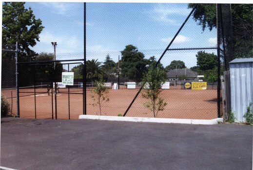 Tennis courts surrounded by a high black wire and pole fence with an asphalt area and shed in the foreground.  Lots of trees around other sides of the fence line.