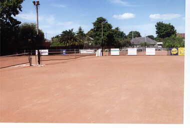 Several tennis courts of red surfaces in front of and behind a metal pole and wire fence, with their tennis nets in place and a tall light pole in the foreground.  Lots of trees and some houses behind and to the left side of the courts.