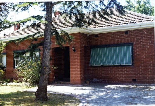 Partial front view of brown brick house with small porch and striped awnings.  Large tree and lawn in the foreground.