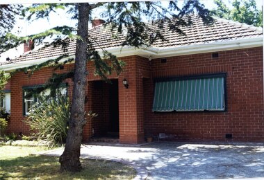 Partial front view of brown brick house with small porch and striped awnings.  Large tree and lawn in the foreground.