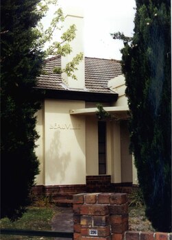 Partial front view of cream house with a tall chimney through tall bushes at the front gate with a path going to the front porch.  The porch has a couple of steps and a circular roof. 