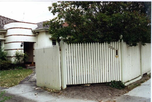 Partial view of a white house from the front gate to the porch with several narrow bands of brown brick.  Most of the foreground shows a tall white picket fence  with a large bushy tree behind it. 