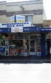 2 storey brick shop and dwelling with many signs and window displays including the sign "MURREMBEENA MILK BAR", with another similar shop to the left and a one storey shop to the right.