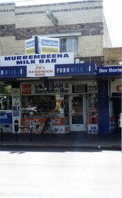 2 storey brick shop and dwelling with many signs and window displays including the sign "MURREMBEENA MILK BAR", with another similar shop to the left and a one storey shop to the right.