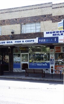 2 storey brick shop and dwelling with several signs including the sign "BLUE SEA FISH & CHIPS", with other similar shops to either side.  Large street seat on the footpath in front of the shop.