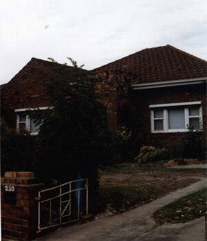 Partial view of brown brick house and matching low fence.  It has a porch with 2 arches, white framed windows, rolled up striped awnings, a white metalwork gate and broken up lawn to the left of the drive.