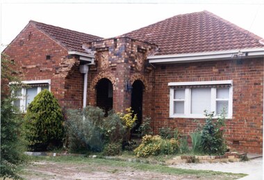 Partial view of brown brick house a porch with 2 arches, white framed windows, rolled up striped awnings, a white metalwork gate and broken up lawn to the left of the drive.  Brickwork patterns stand out in this closer view.