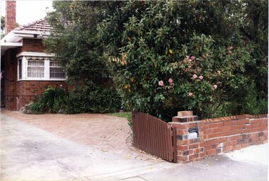 Partial view of a brown brick house showing its left side with tall chimney and white windows from the front gate.  Most of it is hidden by bushes and trees.