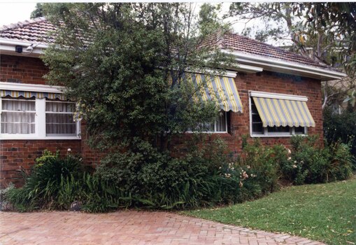 Partial front view of a brown brick house with 3 sets of white windows with striped awnings.  In front of it are curved garden beds of well-established plants and a paved area to the left.