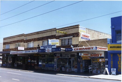 A group of 5 double two-tone brown brick storey shops and one single storey light brown shop, all with awnings over the footpath. Portion of a two storey blue shop at the right end also visible. 