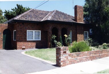 Front view from the left side drive of a mixed browns brick house with one rectangular entry to the porch which also has two large open arches.  Side gate and wall (same bricks) attached to the left side of the house and a brick fence with a few established plants in the front garden.