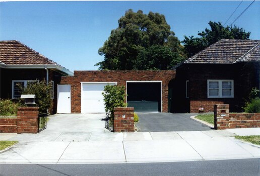 Joined garages of matching mixed browns bricks for 2 different neighbouring houses which are also joined to their garages.  White garage door and rectangular side door on left side and dark garage door and arched side door on the right, with similar but different matching brick fences and drives.