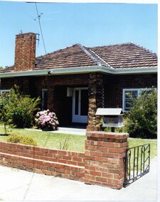 Partial front view of a mixed brown brick house with an open porch including brick pillar and white trellis on the house wall, a wide tall feature brick chimney plus white-framed front door, windows and guttering. The established garden is behind a low brown brick fence with a timber letterbox on the gate pillar and a black metal-work gate.