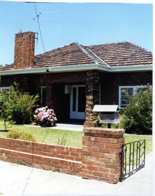 Partial front view of a mixed brown brick house with an open porch including brick pillar and white trellis on the house wall, a wide tall feature brick chimney plus white-framed front door, windows and guttering. The established garden is behind a low brown brick fence with a timber letterbox on the gate pillar and a black metal-work gate.