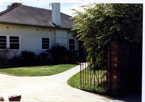 Partial view of front of white house with a tall chimney, white windows and front path. The foreground has a black metal-work gate and tall brown brick pillar alongside the established garden.