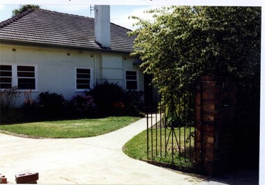 Partial view of front of white house with a tall chimney, white windows and front path. The foreground has a black metal-work gate and tall brown brick pillar alongside the established garden.