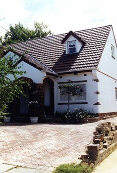 Partial view of front and side of a double storey white house with steep roof which has a little roofed window jutting out of it. Brown brick decoration including around the double arched porch.  Paved drive and front area.