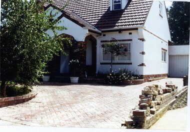 Partial view of front and side of a double storey white house and its garage.  It has a steep roof with a little roofed window jutting out of it. Brown brick decoration including around the double arched porch. Paved drive and front area with bricked garden beds.