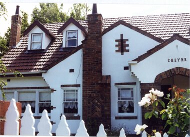Partial view of front of a double storey white house with steep roof which has 2 little roofed windows jutting out of it. Brown brick decoration including around the arched porch, windows and chimneys. Part of a tall white picket fence and garden visible in front of the house.