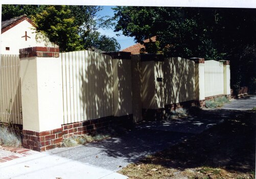 High brick and timber paling fence, cream and brown in colour, with a gate 1/3 of the way, which overlooks a street footpath.  There is a similarly coloured building behind the left corner pillar with tall trees behind that, and a tiled red roof to the right.