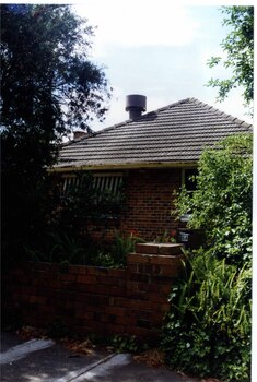 Brown brick fence with a letterbox and side of a matching brown brick house with a window and its half-open striped window blind.  House is partly hidden by trees and bushes in the garden.