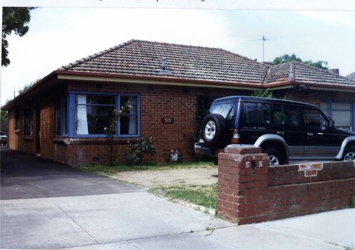 Side and front of brown brick house which has a brick front fence to the right of the drive going down the side of the house.  Windows have blue frames.  Big black and grey car on the front lawn with a few plants in the garden.