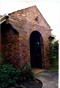 Close view of decorative arched brick porch with a metal lantern above the arch and a window on the right wall of the porch.  Garden beds each side with garden path to the porch.